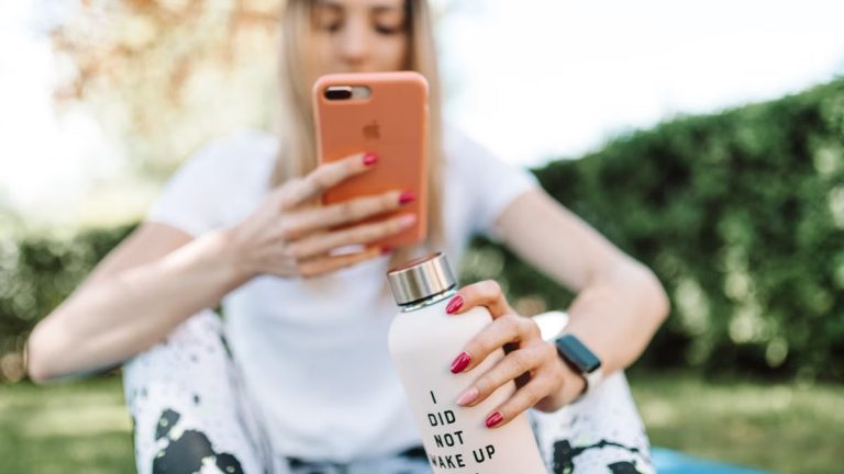 Woman outdoors using smartphone while holding a water bottle with text. Casual and relaxed setting.